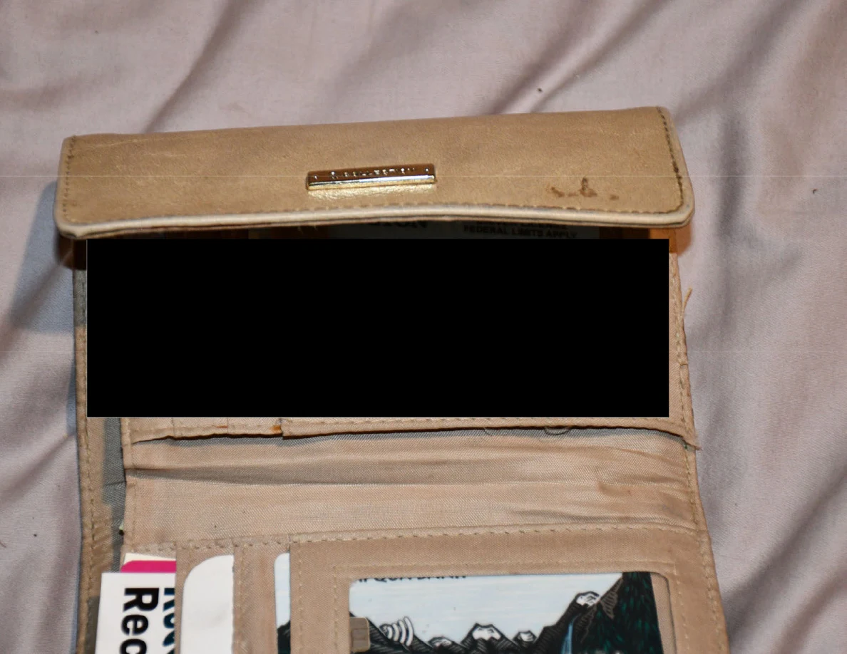 Open tan wallet on bedding with its contents partly obscured by a black redaction.