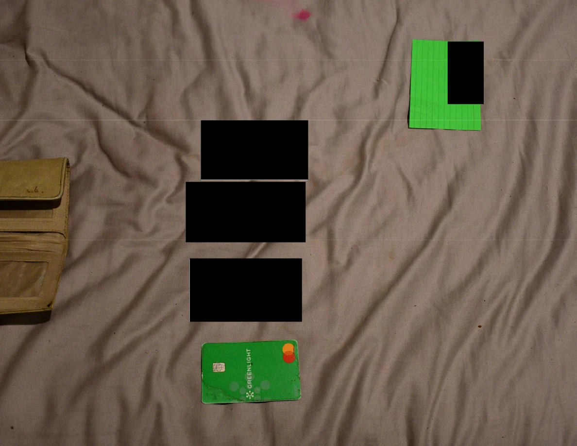 Open wallet and a row of partly redacted cards on bedding beside a green note.