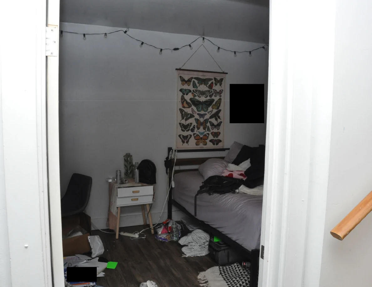 View through a bedroom doorway showing a bed, butterfly wall hanging, string lights, bedside drawers and scattered floor items.