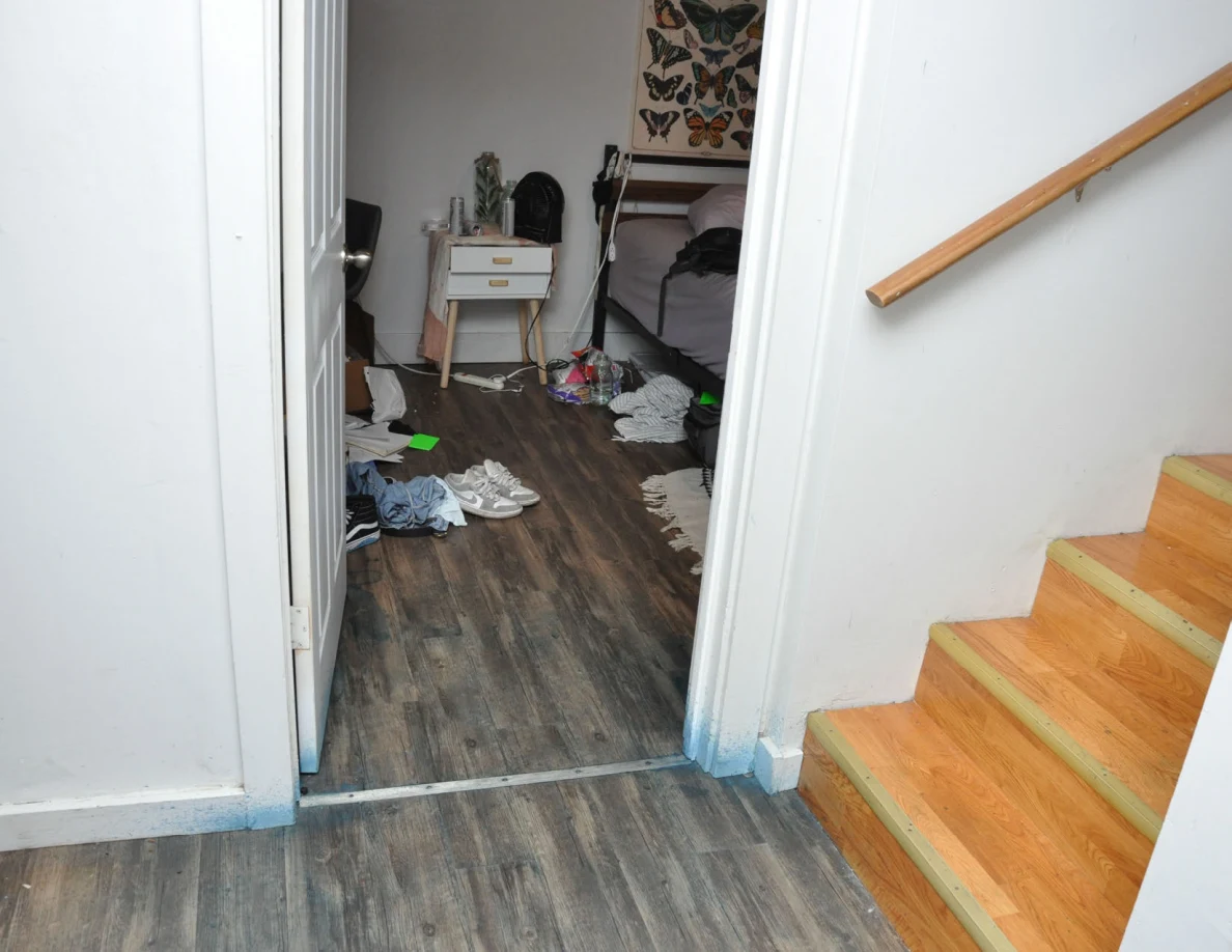 Bedroom doorway beside wooden stairs, with shoes, scattered items and bedside drawers beyond.