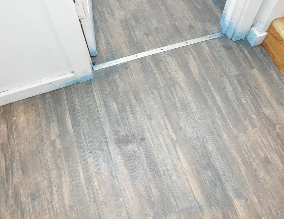 Wood-pattern floor and metal bedroom threshold beside stairs, with blue discoloration at the door trim.