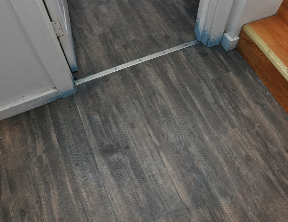 Angled view of a doorway threshold and wood-pattern floor beside the bottom stair.