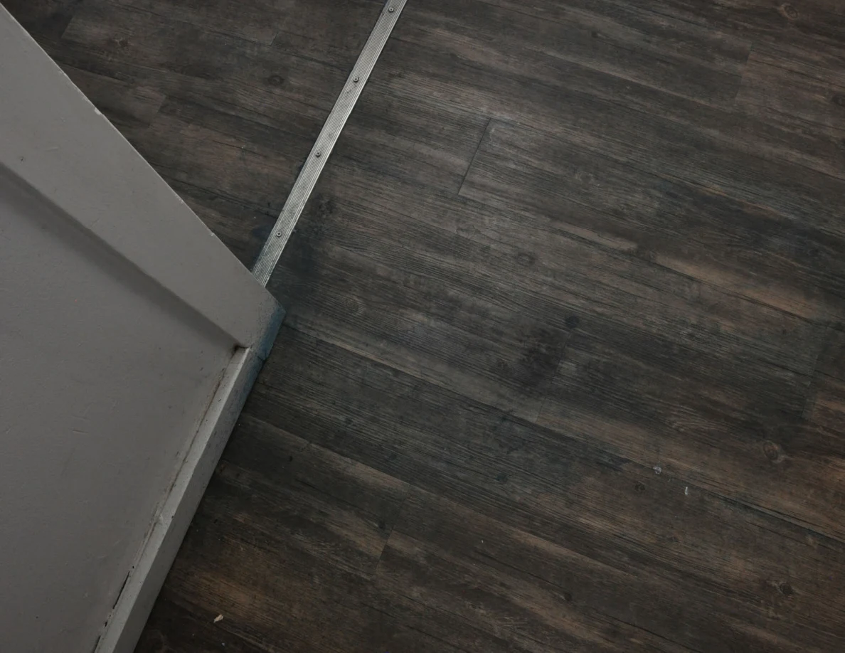 Downward view of wood-pattern flooring and a metal threshold beside a white door frame.