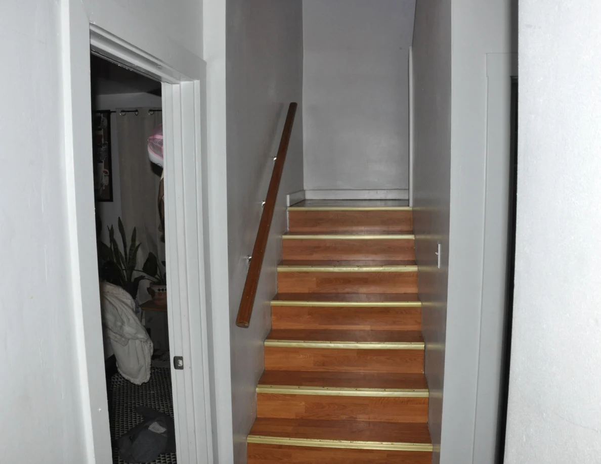 Wooden stairs with a handrail between white walls beside a bedroom doorway.