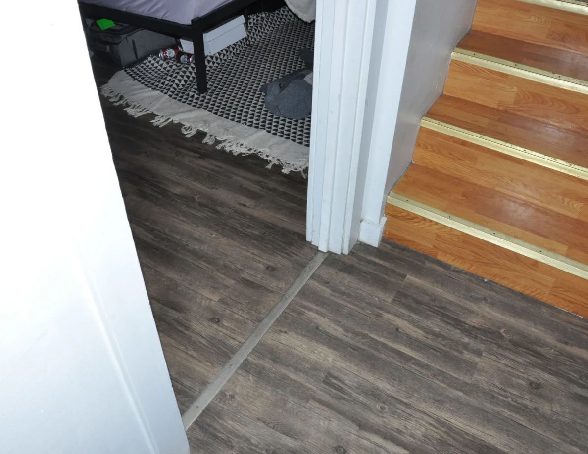 Wood-pattern floor at a bedroom doorway beside wooden stairs and the edge of a patterned rug.