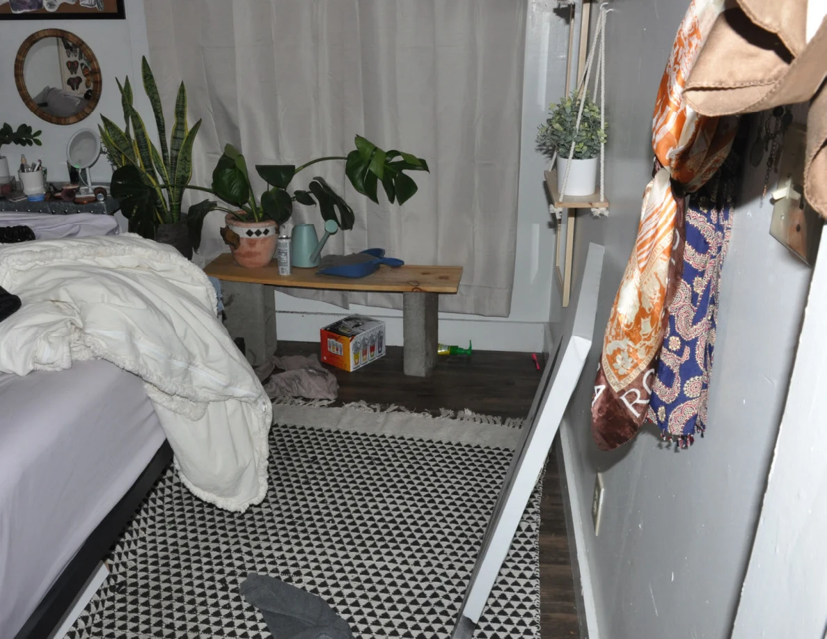 Bedroom corner with potted plants on a wooden bench, curtains, hanging bags and the edge of a bed.