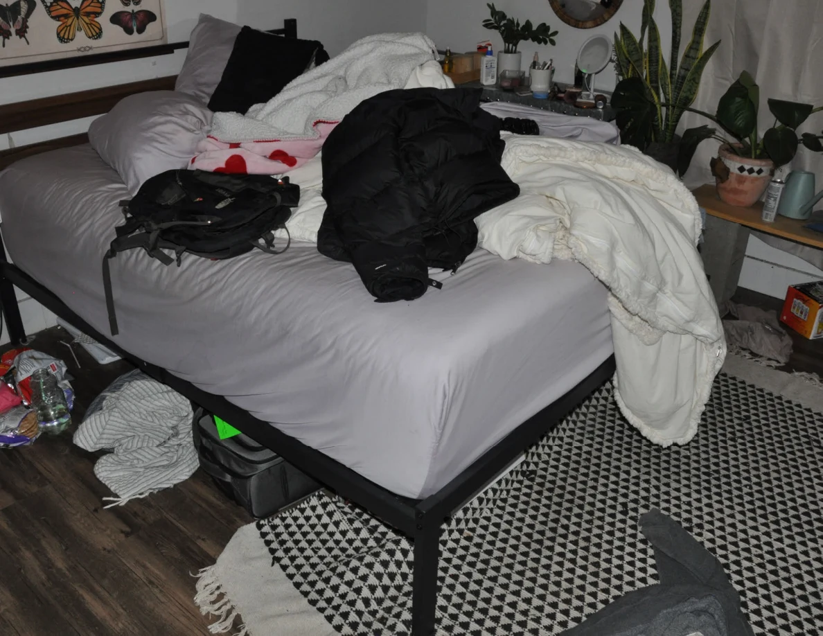 Bed with pale bedding, dark clothing and a black bag above a patterned rug.