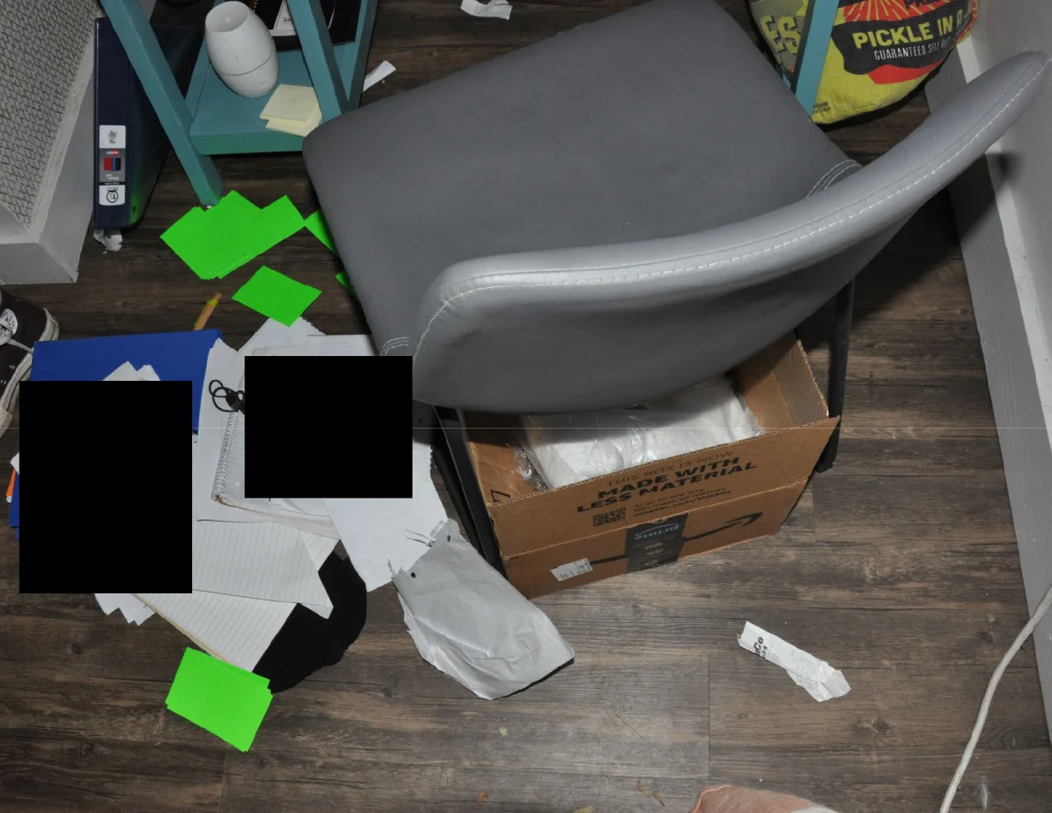Gray chair over a cardboard box beside scattered papers, green markers and loose packaging on the floor.