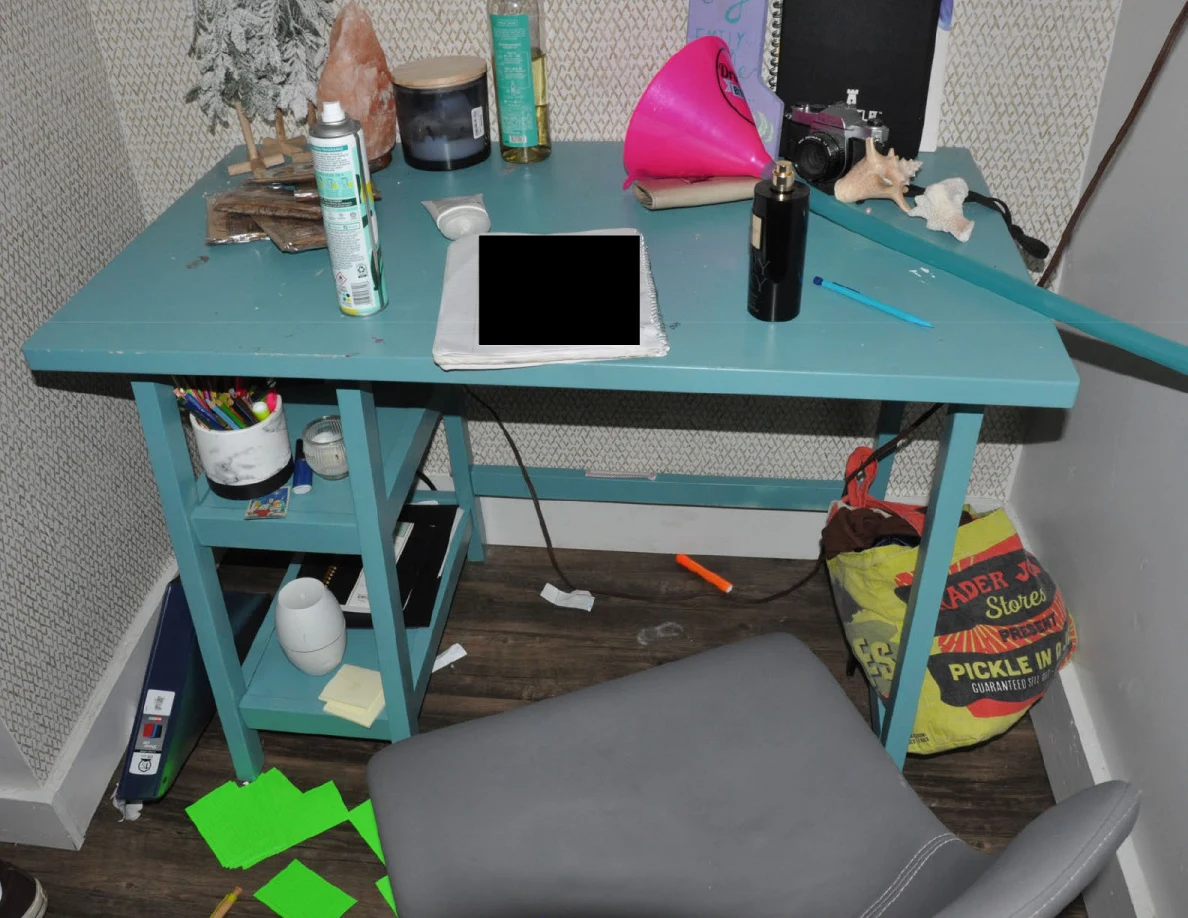 Turquoise desk with bottles, toiletries, pens, small objects and a redacted notebook beside a gray chair.