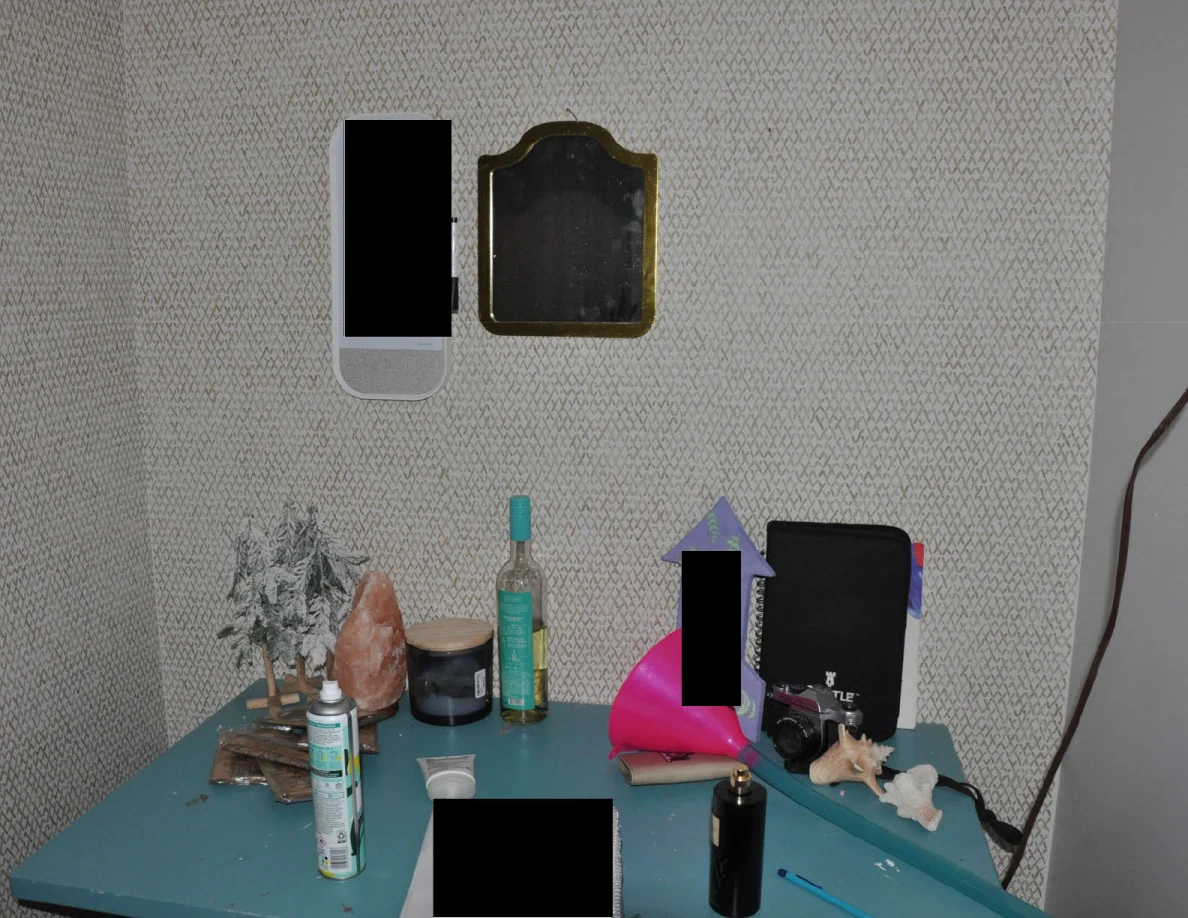 Turquoise desk beneath small wall mirrors with bottles, toiletries, a pink pouch and redacted items.
