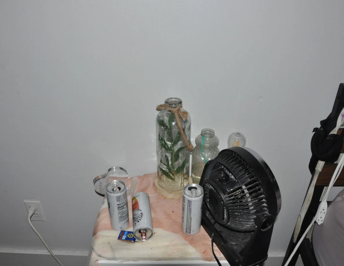 White bedside table holding a black fan, drink cans and glass containers with plant stems.