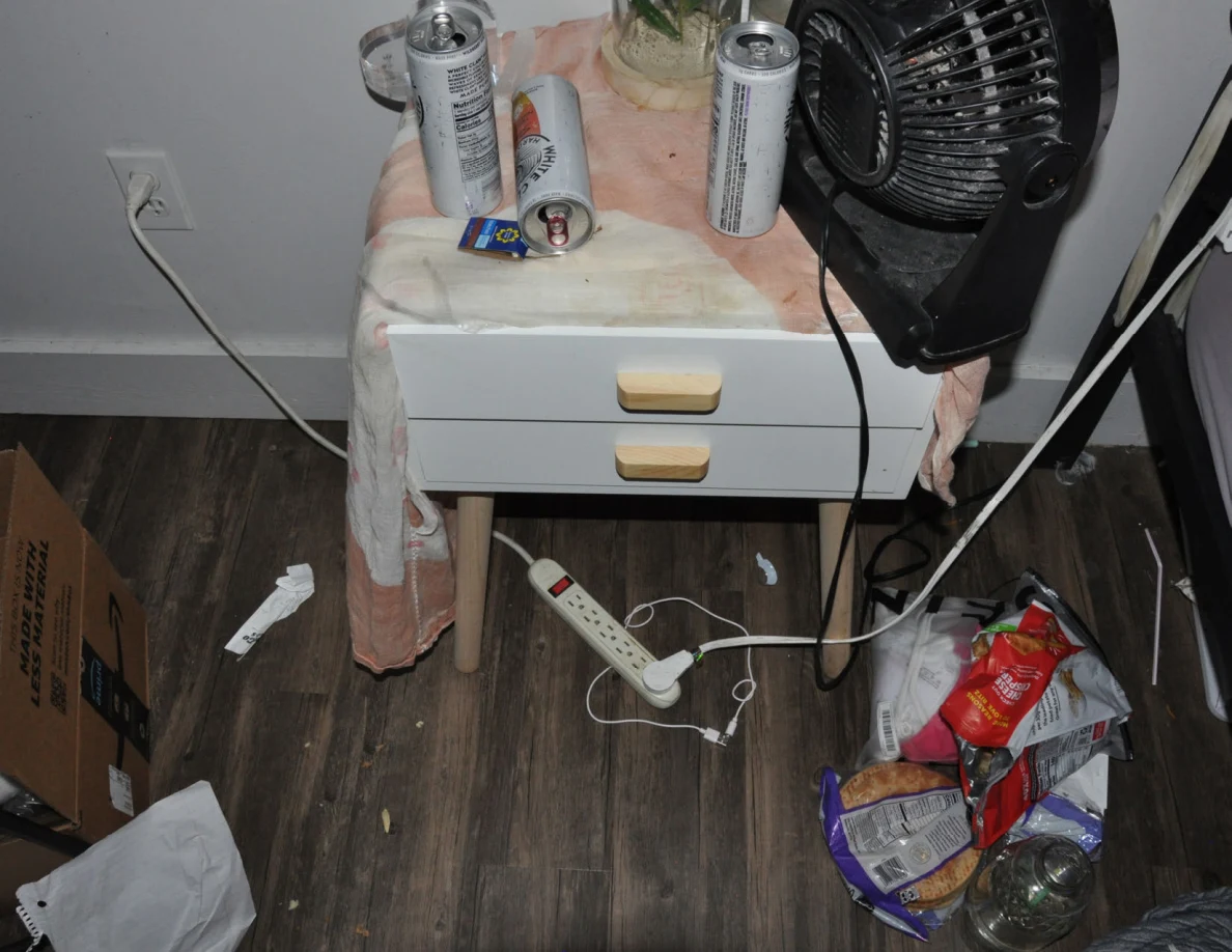 Bedside table with fan and drink cans above a power strip, cables and snack packaging on the floor.