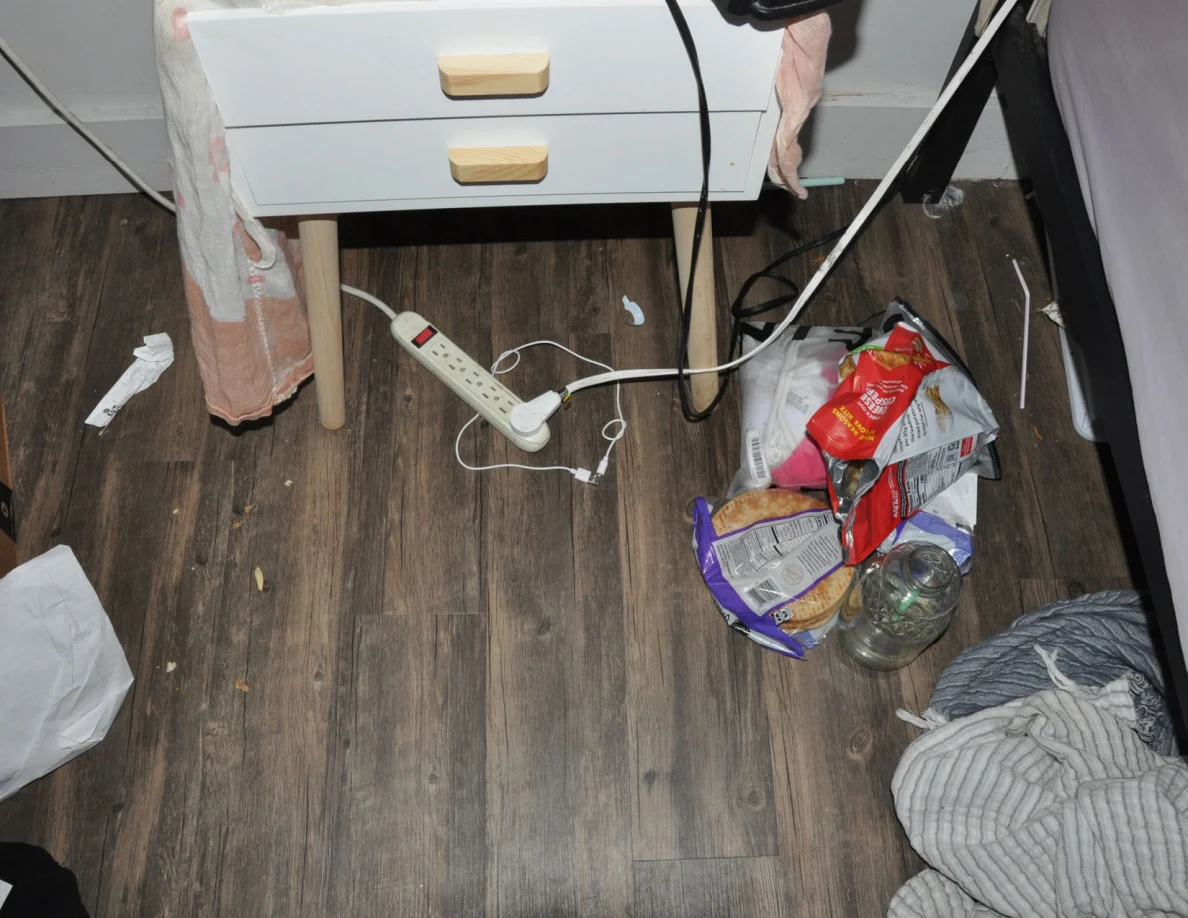 Floor beside a bedside table with a power strip, cables, snack bags, glass container and loose clothing.