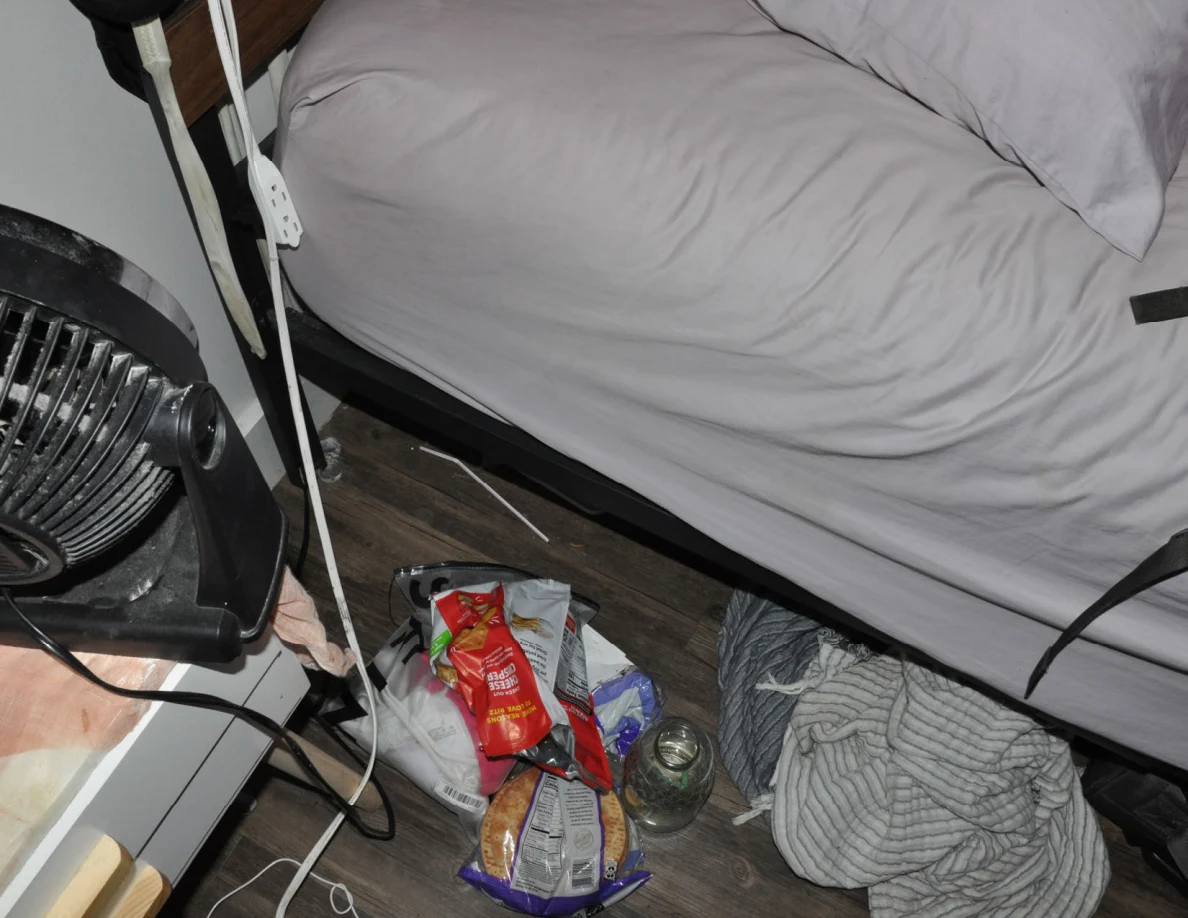 Side of a bed above floor snack bags, a glass jar and loose striped bedding beside a fan.