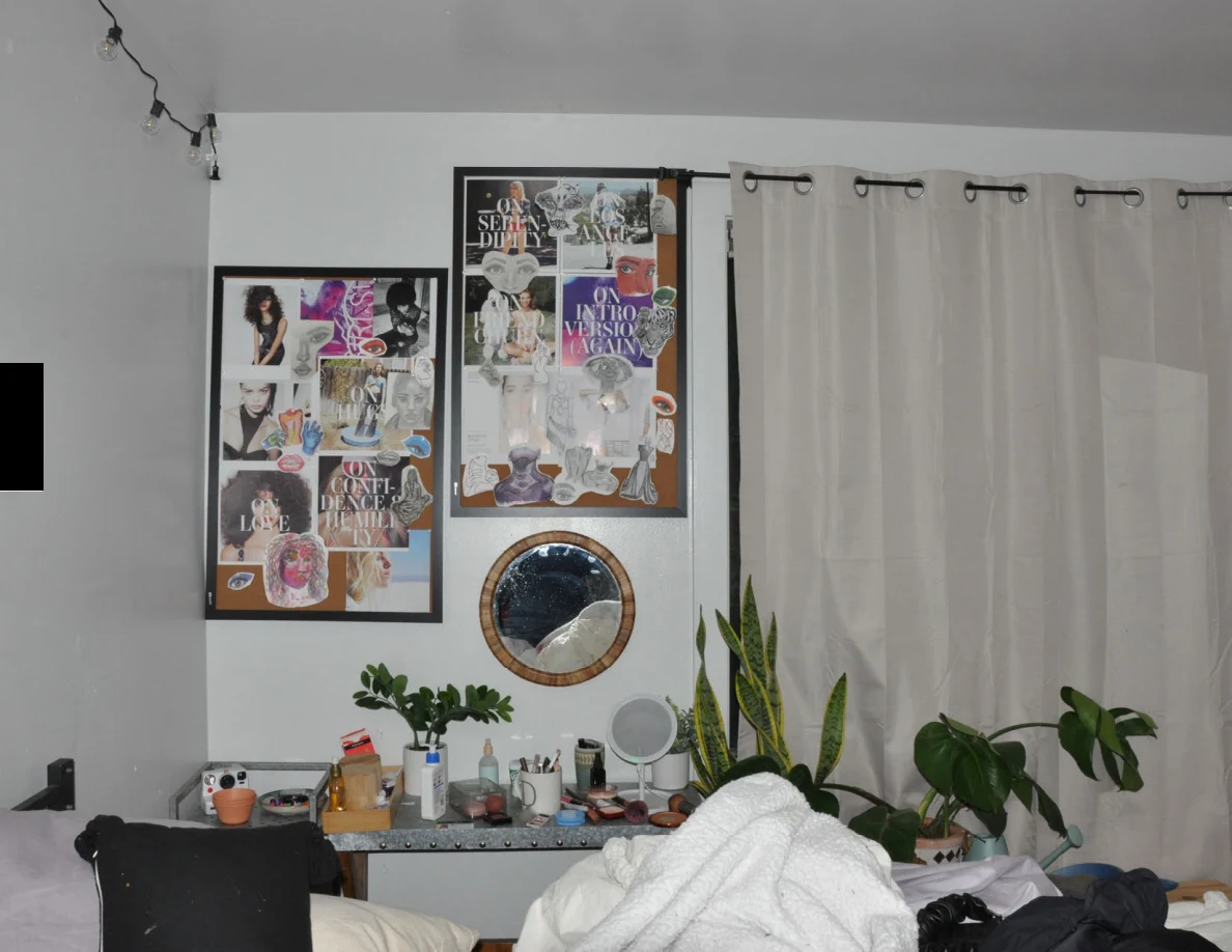 Framed wall collages and round mirror above toiletries and potted plants beside closed curtains.