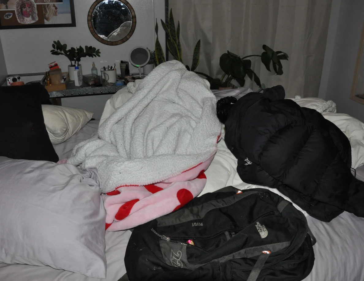 Rumpled bed with a white blanket, pink patterned blanket, black jacket and black backpack.