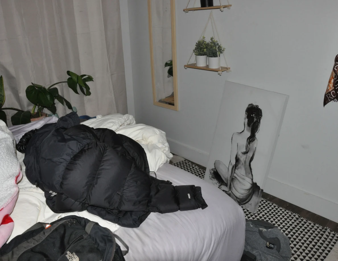 Black jacket on a bed beside a wall mirror, hanging plants and a drawing leaning against the wall.