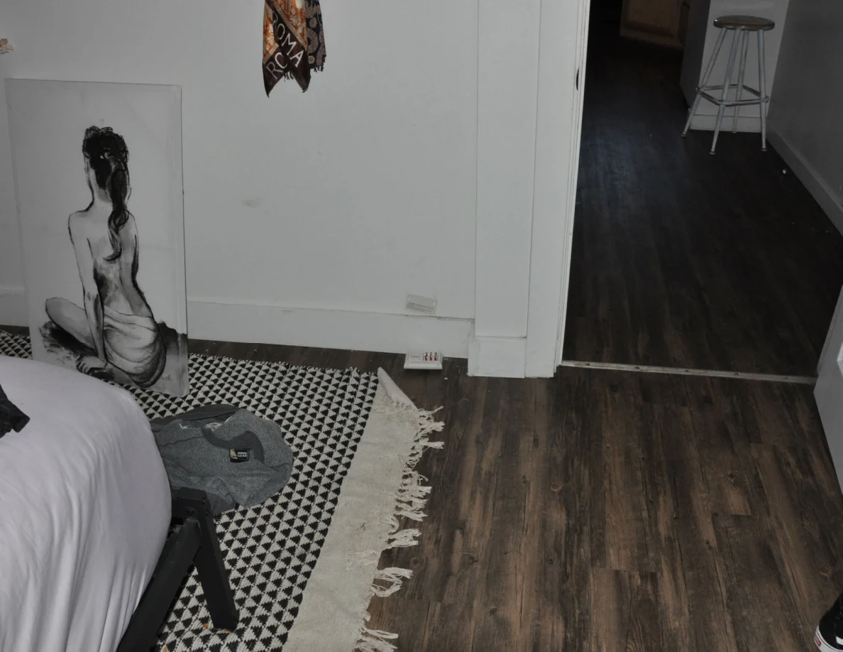 Bedroom floor and patterned rug beside a leaning figure drawing and open doorway toward a stool.