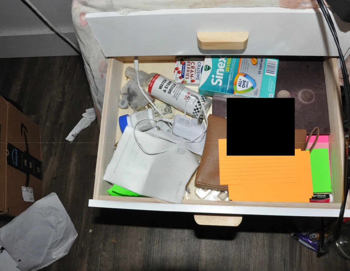 Open bedside drawer with medicine packaging, cables, colored note cards and a partly redacted item.