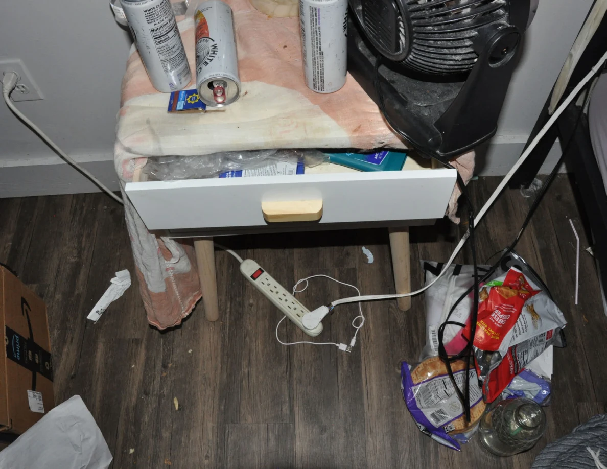 Partly open bedside drawer beneath drink cans and a fan, with cables and packaging below.