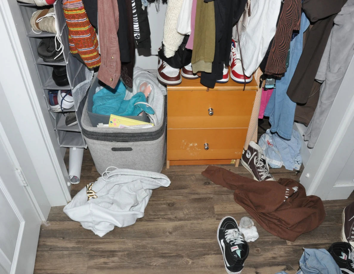 Open closet with hanging clothes, wooden drawers, gray storage basket and loose clothing and shoes.