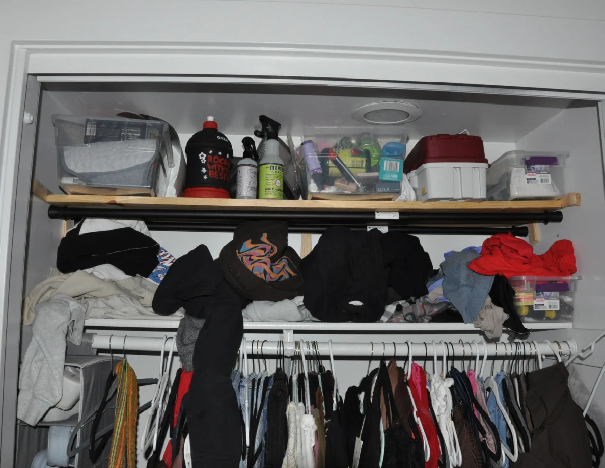 Upper closet shelves with storage containers, bottles and piled clothing above hanging garments.