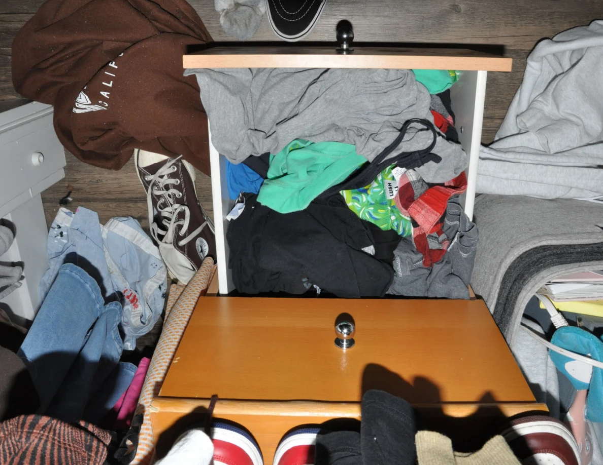 Open wooden drawer filled with mixed clothing beneath a closet rail.