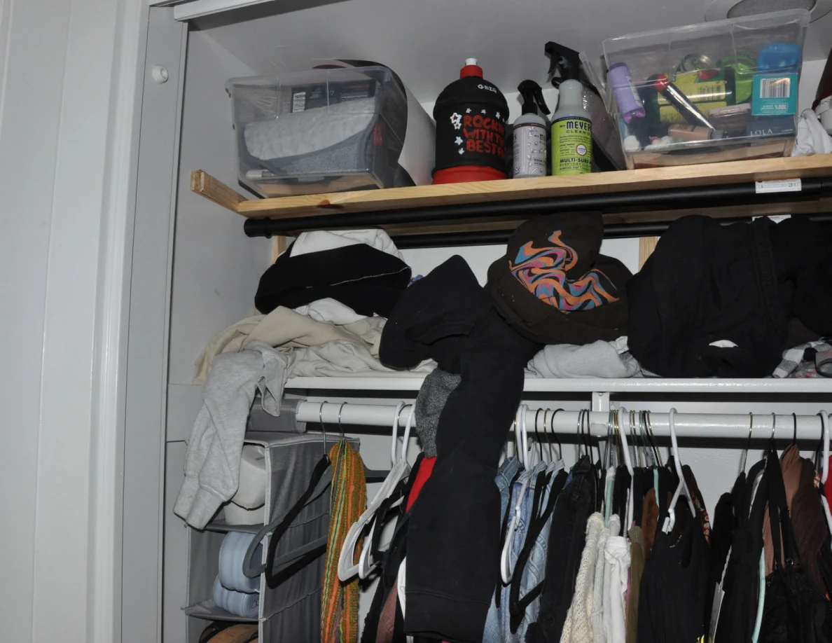 Closet shelves with containers, bottles and piled clothing above a packed clothes rail.
