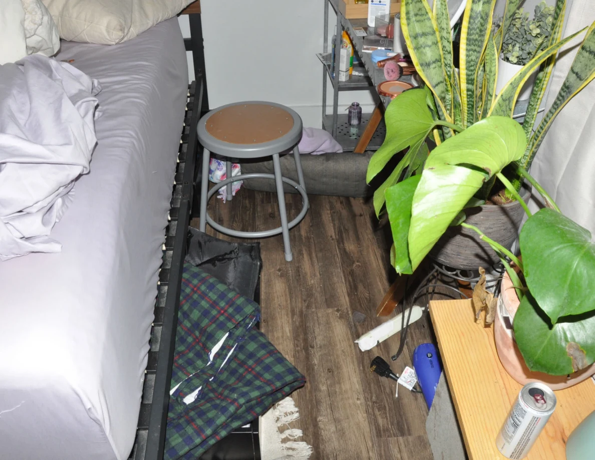 Narrow floor space beside a bed, stool and plants with plaid fabric and small scattered items.