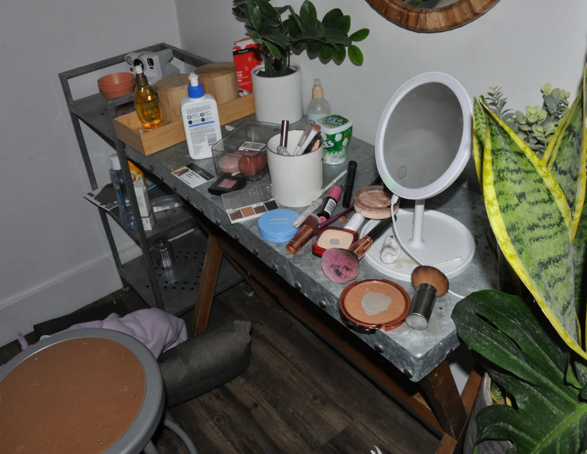 Cosmetics table with makeup brushes, bottles and a round tabletop mirror beside plants.