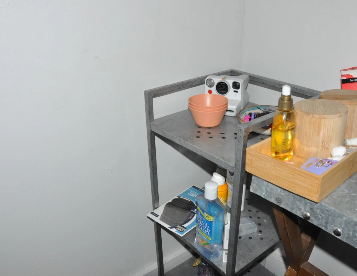 Metal shelving beside a cosmetics table holding an instant camera, small bowl and bottles.