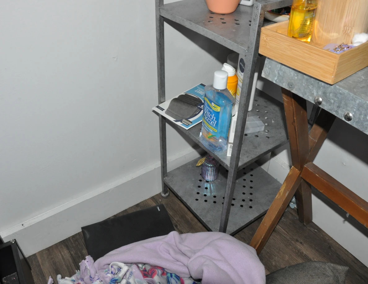 Lower metal shelving with bottles and paper items beside a table and loose bedding.