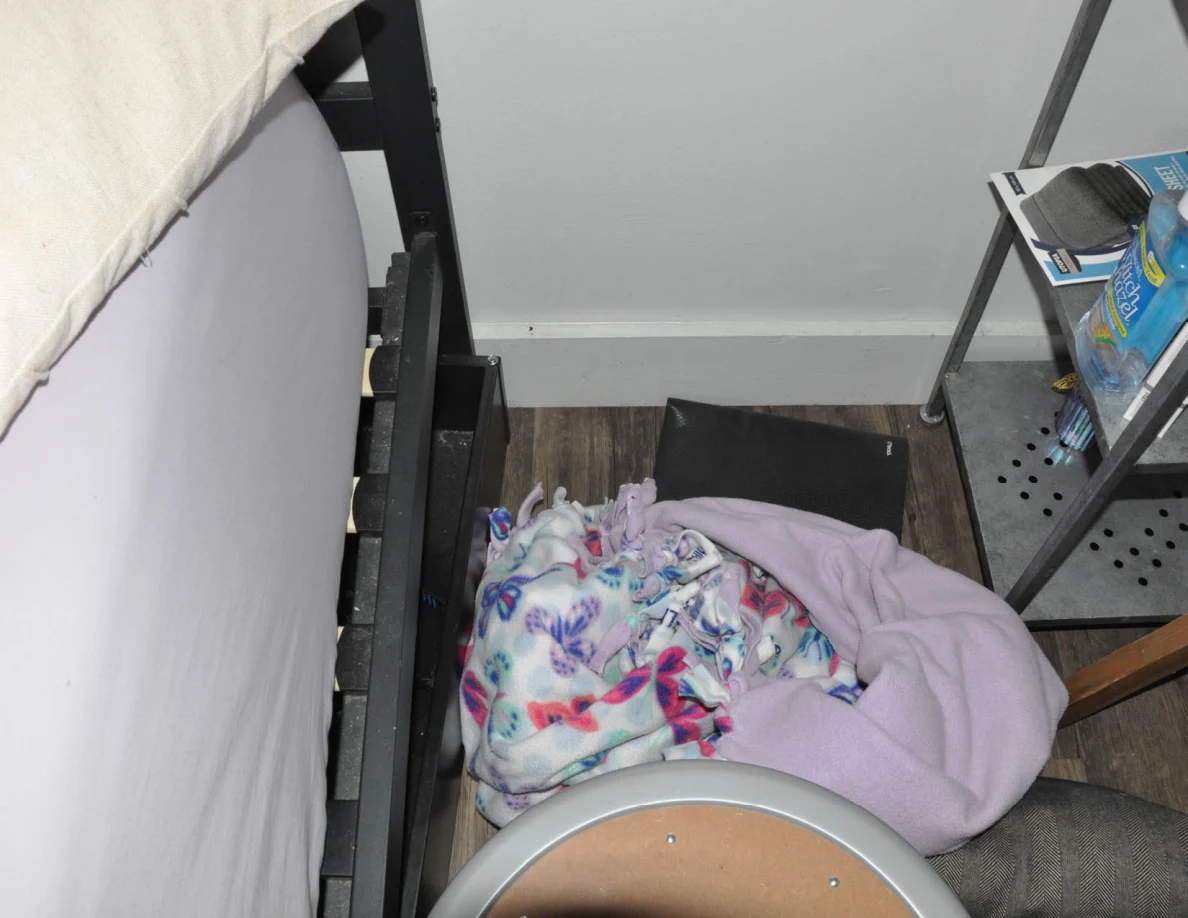 Floor between a bed and shelves with colorful fabric and a pale purple blanket.