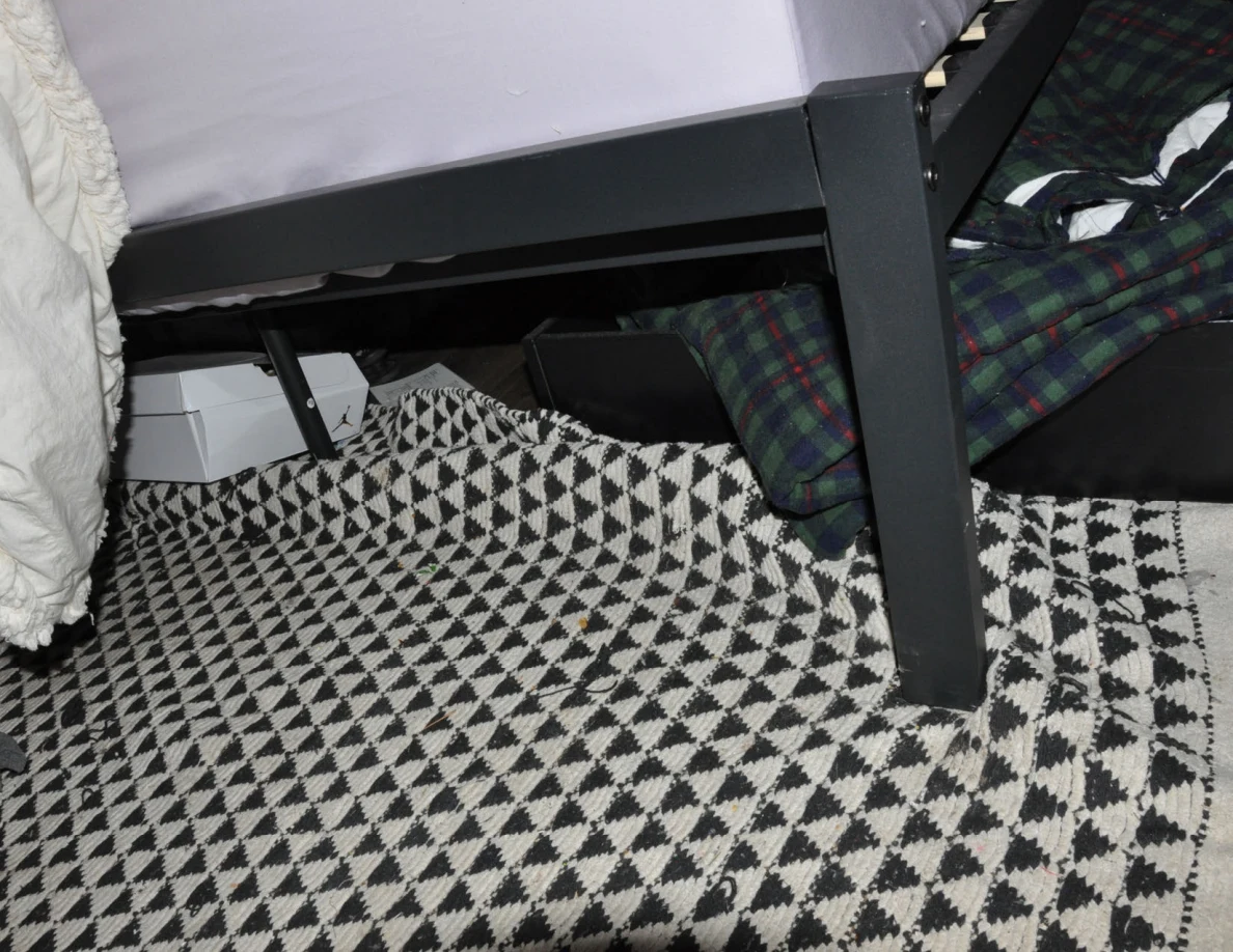 Bed leg and black-and-white patterned rug with boxes and plaid fabric beneath the bed.