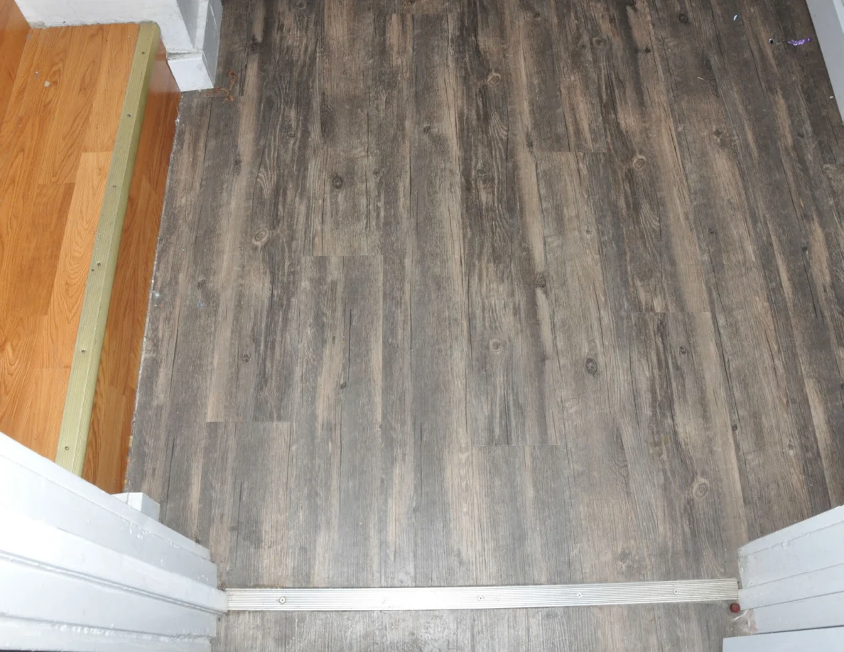 Downward view across a metal doorway threshold to wood-pattern hall flooring beside a wooden step.