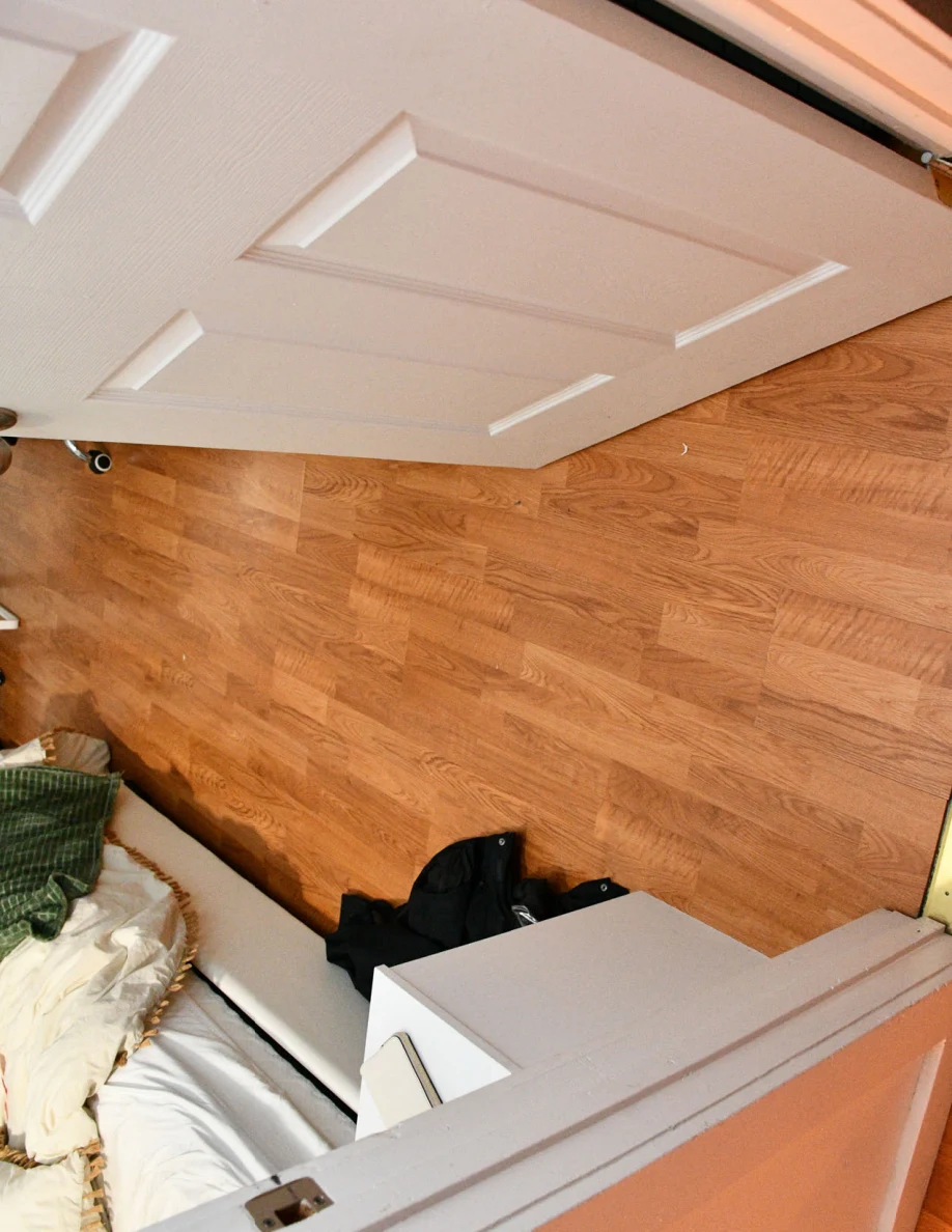 Downward view through an open white door to wood-pattern flooring, bed corner and a black bag.
