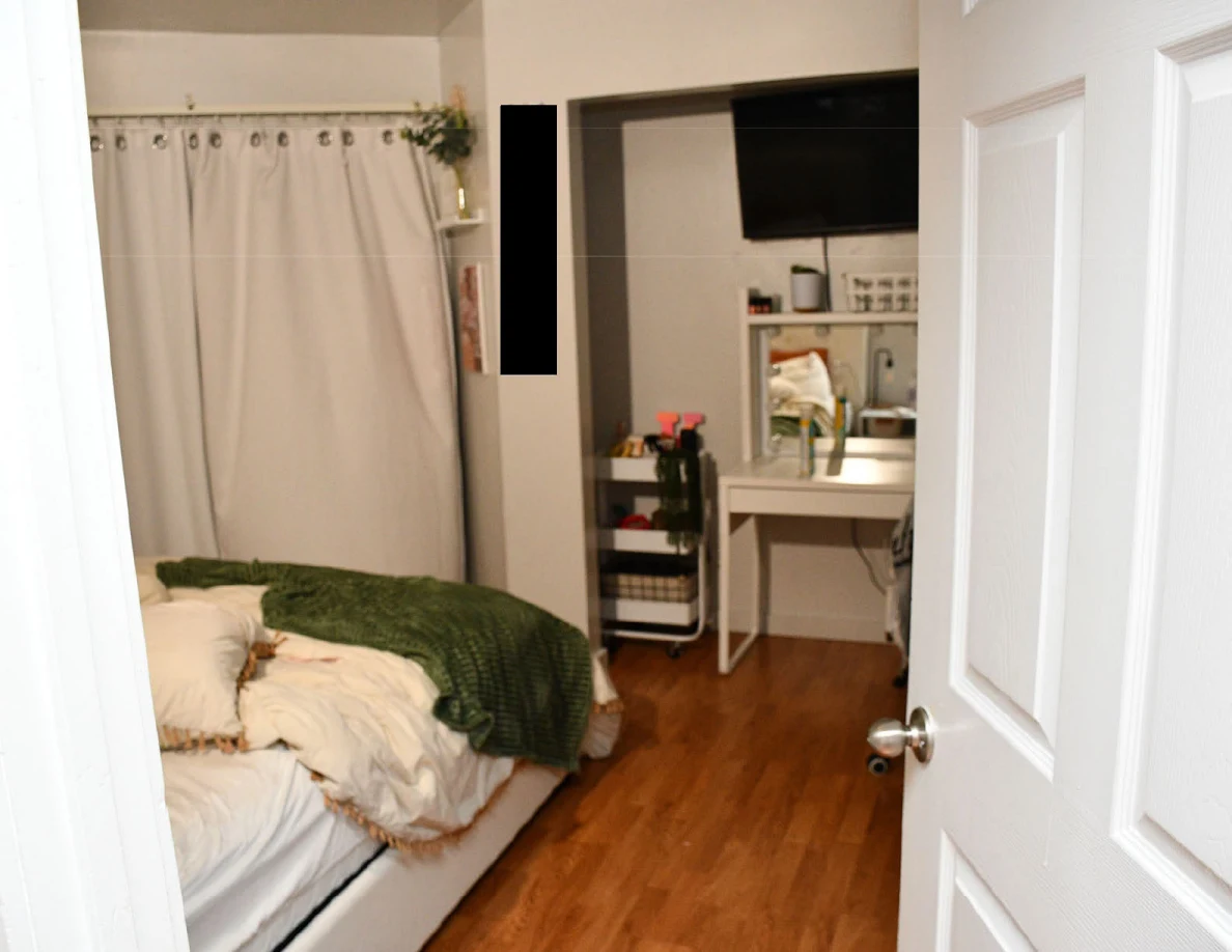 Bedroom seen through an open door with white bedding, green blanket, curtains and a white vanity beneath a television.