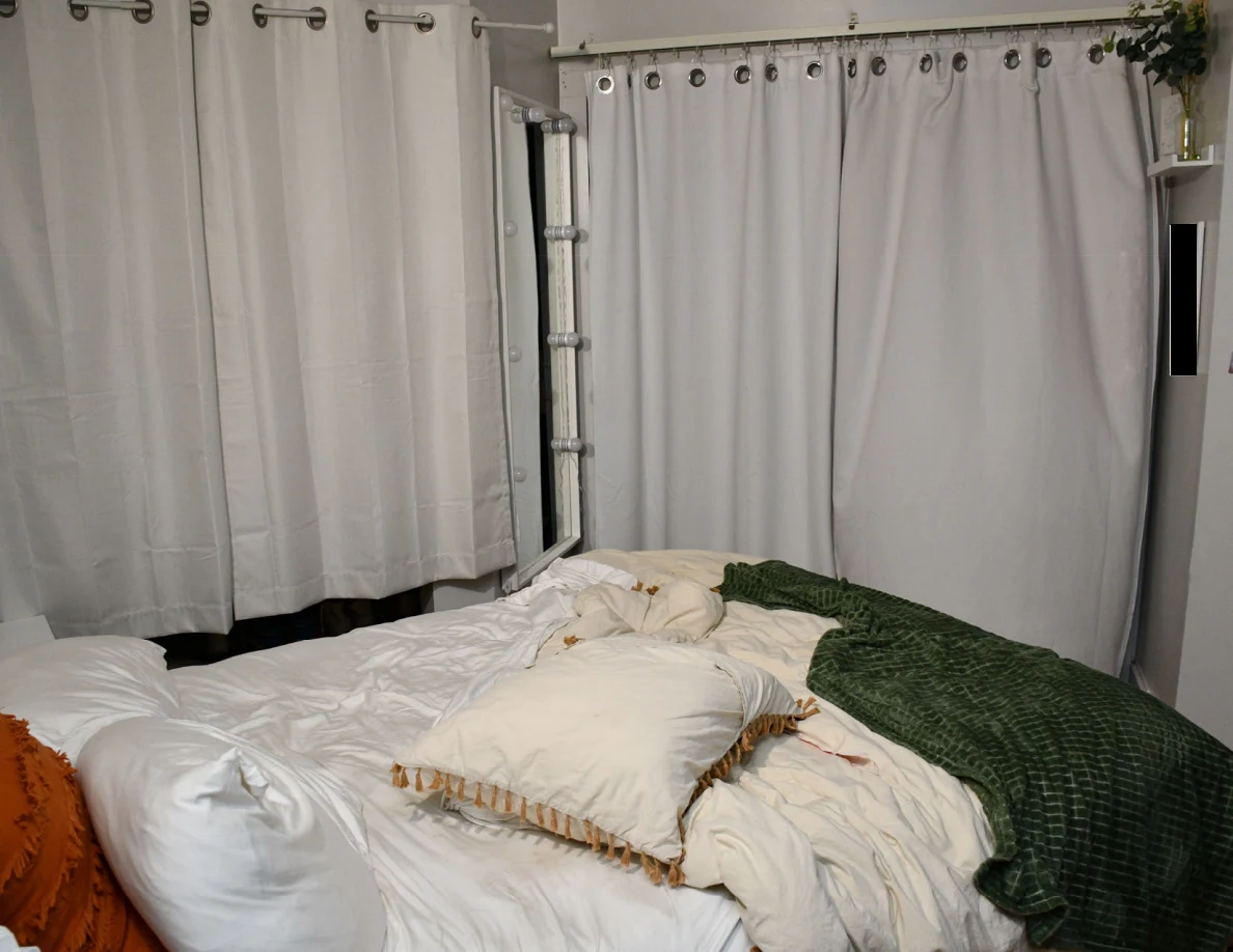 Rumpled white bedding and a green blanket beneath curtains with a tall mirror in the corner.