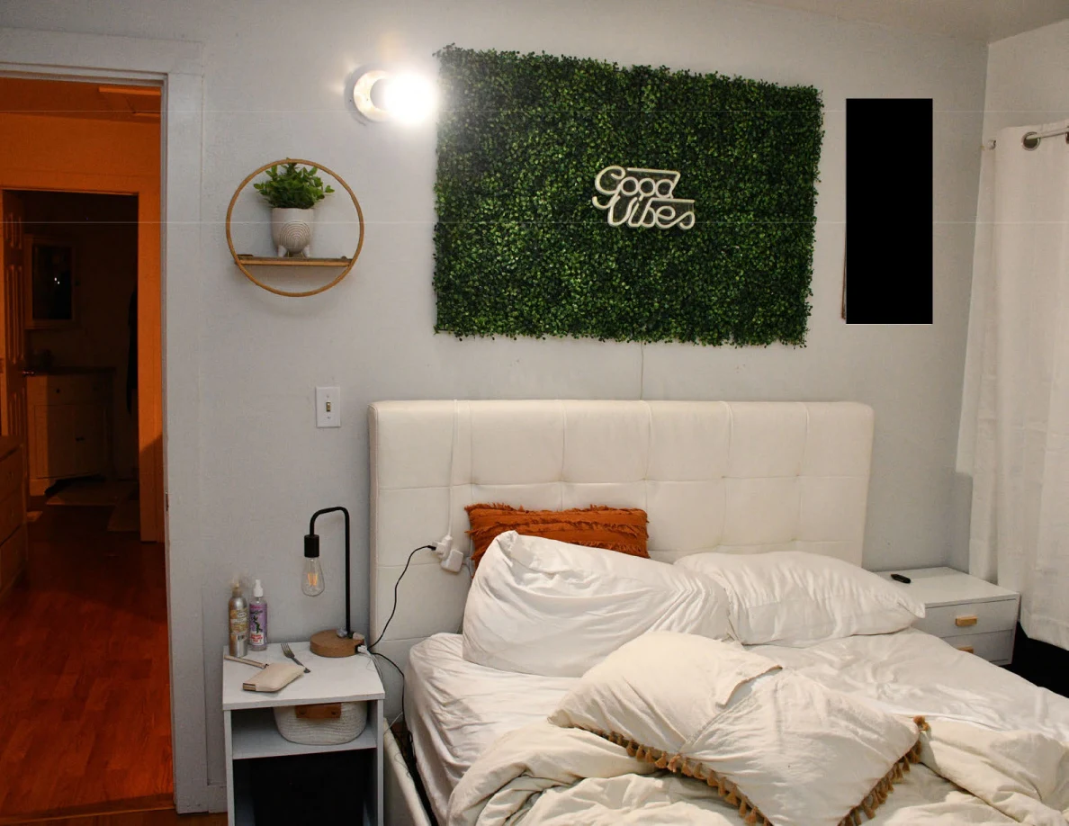 Bed beneath an artificial greenery panel reading Good Vibes, with bedside tables and a wall planter.