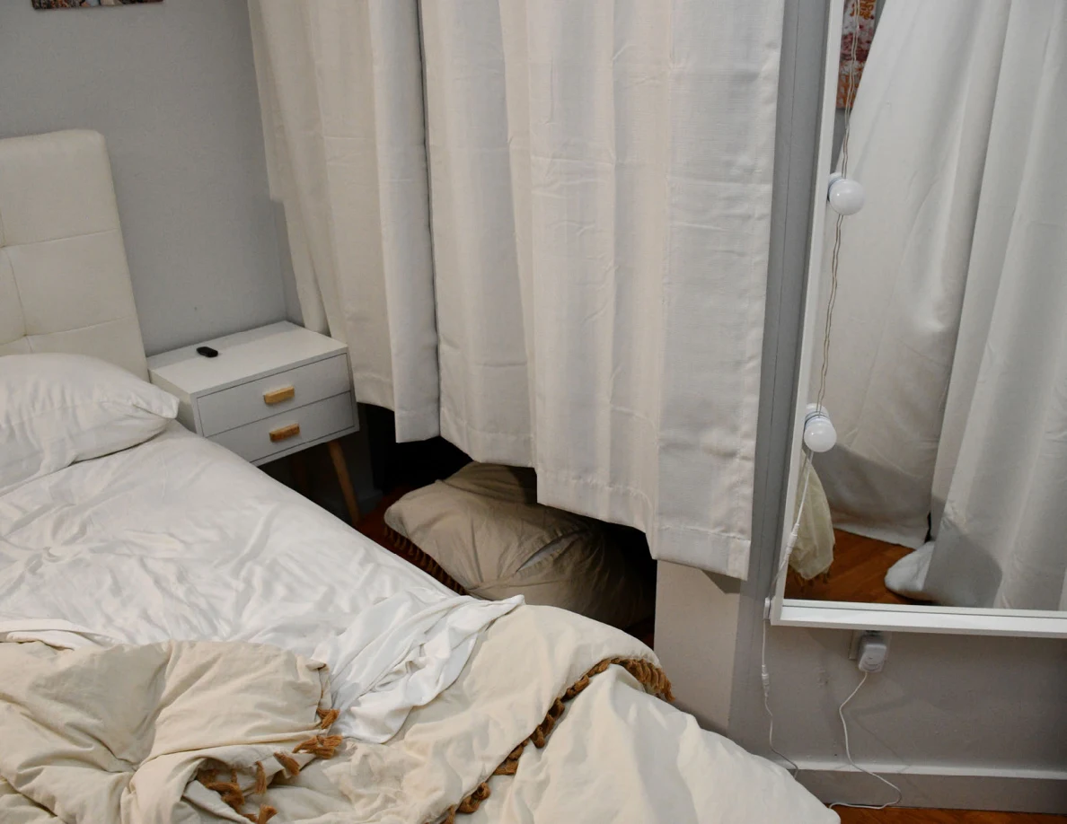 Bedside table and rumpled bedding beside curtains and a tall bulb-edged mirror.