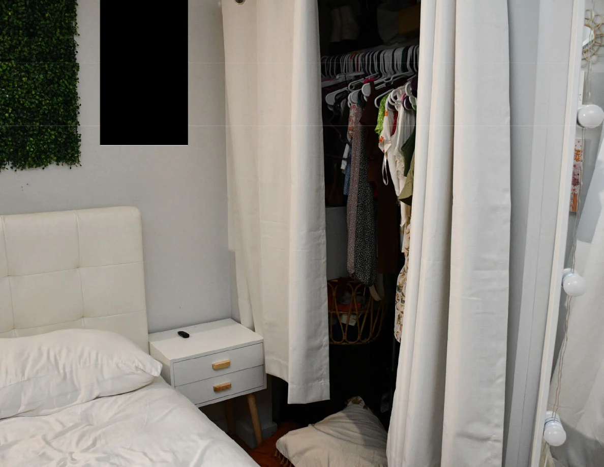 Partly opened curtain revealing clothes in a closet beside a bed and mirror.