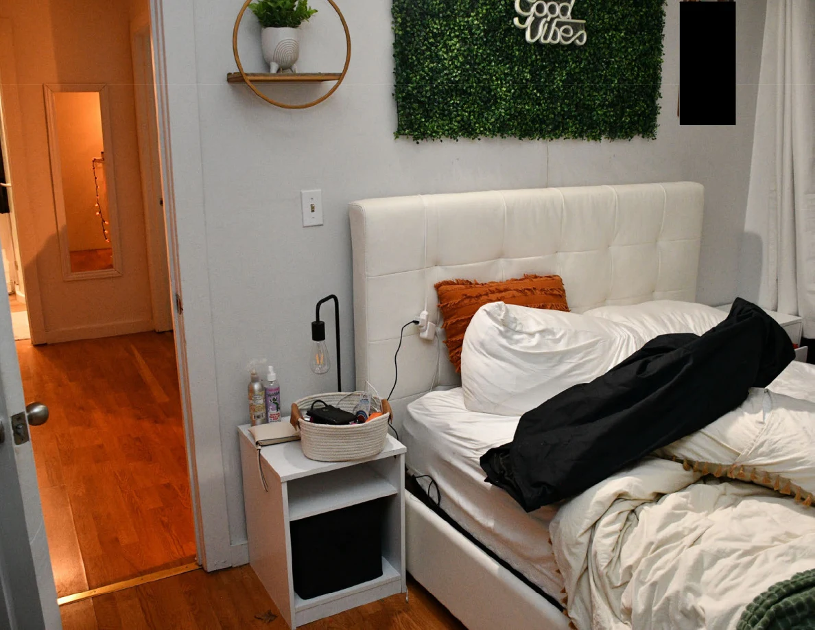 White headboard and bedside table with a woven basket beside a black fabric bag on the bed.