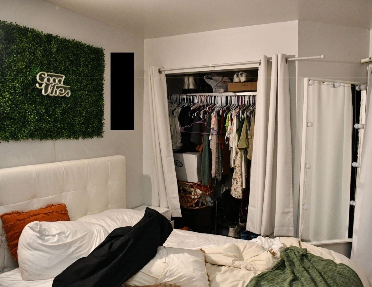 Bedroom with open clothes closet and a black fabric bag across rumpled bedding beneath a greenery panel.
