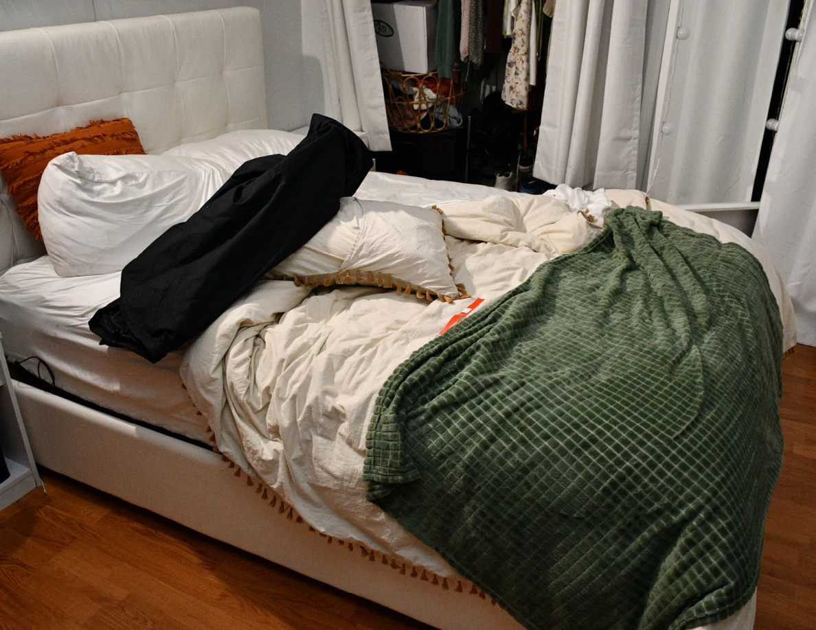 Bed with a green blanket, white pillows and black fabric bag beside an open closet.