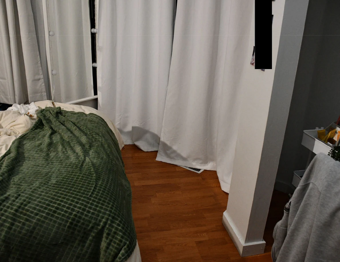 Wooden floor beside the foot of a bed, green blanket and closed white curtains.