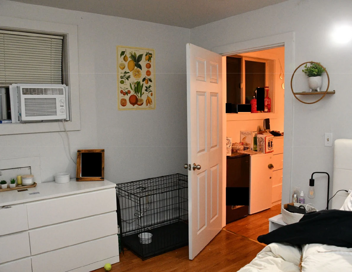 Bedroom window air conditioner, dresser and wire pet crate beside an open orange-lit doorway.