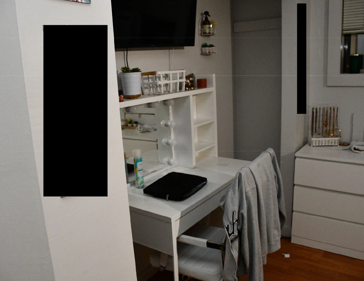White vanity and television beside a dresser and garment-draped chair, with several redacted areas.