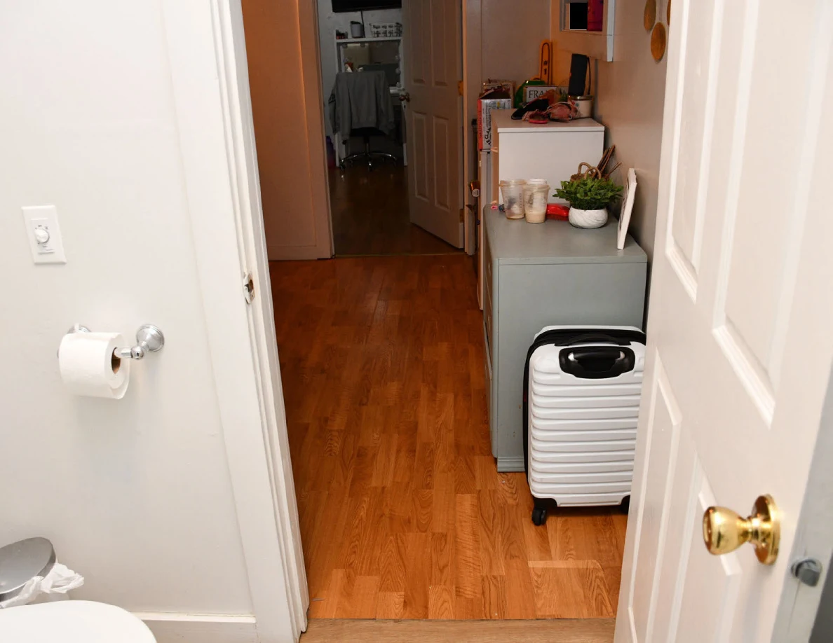 View from a bathroom past a toilet-paper holder into a hall with a white suitcase and small cabinet.