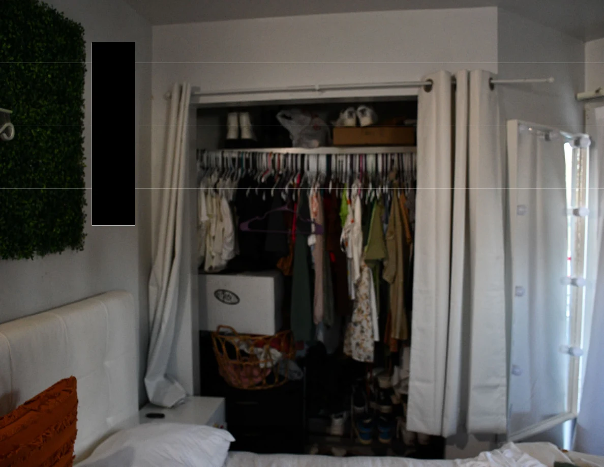 Open curtain-front closet with hanging clothes, shoes, storage boxes and bags beside a bed.
