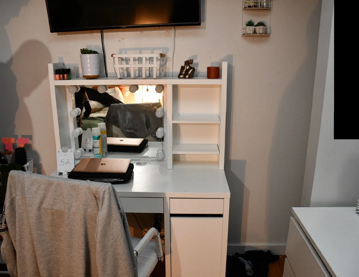 White vanity with a closed laptop and numbered scale card beneath a mirror and television.